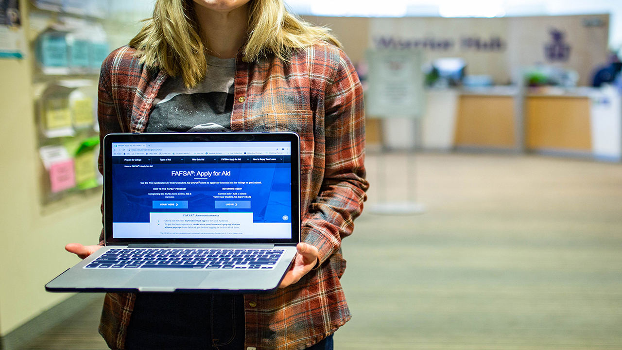 A student in the Warrior Hub lobby holds a laptop showing the FAFSA application 