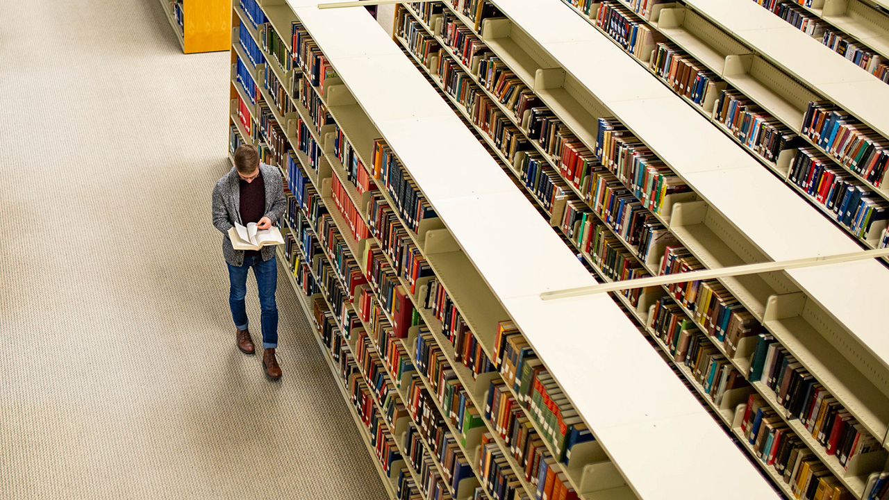 A student walks through the WSU Library while reading a book.