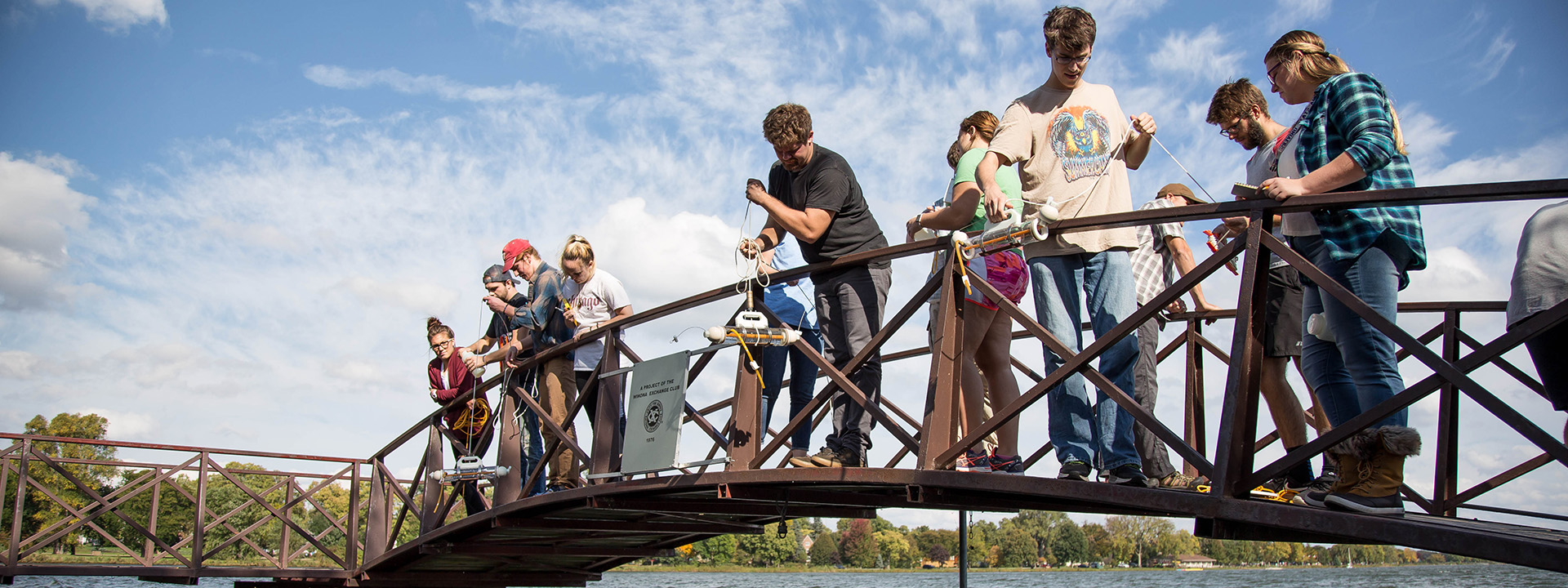 A group of students stand on a bridge over Lake Winona to conduct scientific testing.