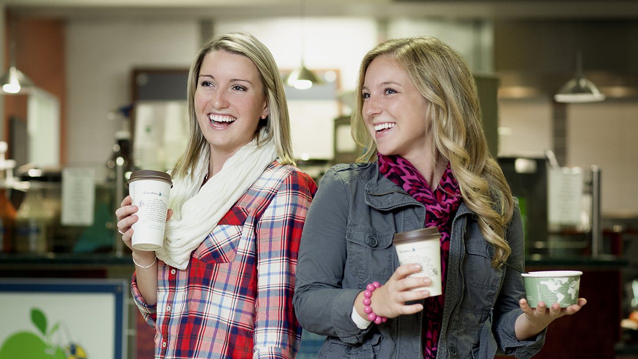 Two female students laugh outside a coffee shop while holding coffee cups. 